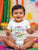 Baby wearing a 'My First Holi' onesie in a photoshoot with colorful decorations in the background.